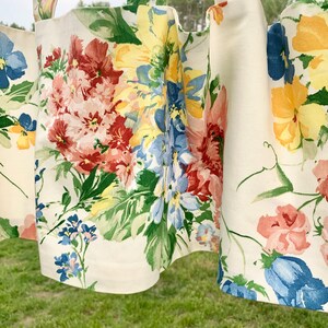 May include: Cream-colored floral curtains with a repeating pattern of large, colorful flowers. The flowers are in shades of red, yellow, blue, and pink, with green leaves. The curtains are hanging outdoors, with a blurred green background.