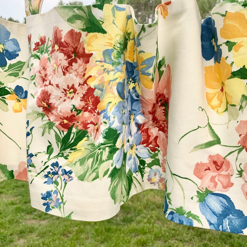 May include: Cream-colored floral curtains with a repeating pattern of large, colorful flowers. The flowers are in shades of red, yellow, blue, and pink, with green leaves. The curtains are hanging outdoors, with a blurred green background.