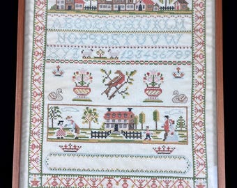 May include: Framed vintage cross-stitch sampler with a wooden frame. The sampler features a detailed stitched design with a village scene, alphabet, numbers, birds, flowers, and figures. The color palette includes red, green, and blue.