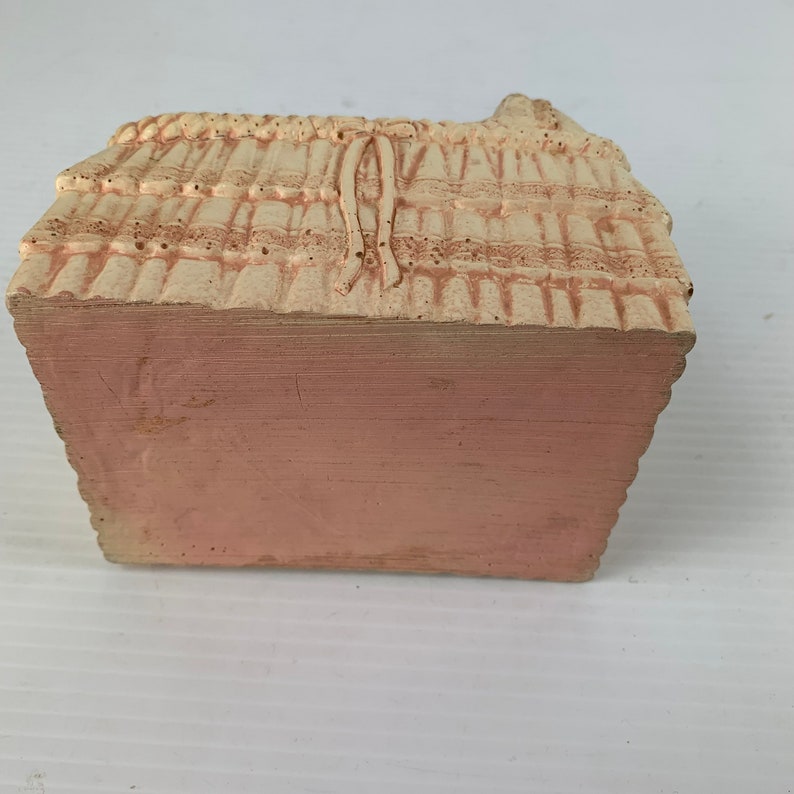 May include: A small, white ceramic basket with a pink base. The basket is decorated with a woven pattern and a bow.