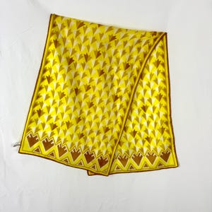 May include: A yellow scarf with a repeating pattern of brown and white arrow shapes. The scarf has a brown border and a decorative brown pattern along the bottom edge. The scarf is folded and lies on a white surface.