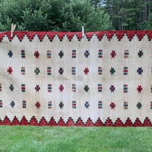 May include: A rectangular, off-white rug with a red border and geometric patterns. The rug is hanging on a clothesline with wooden clothespins. The pattern includes red, green, and blue diamond shapes. The rug has fringed edges and is displayed outdoors.