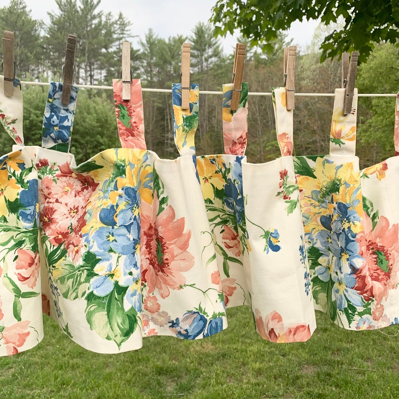 May include: A floral valance with a cream background, featuring large blooms in shades of pink, yellow, and blue. The valance is hung on a clothesline with wooden clothespins. The fabric is visible in detail, showcasing the pattern.