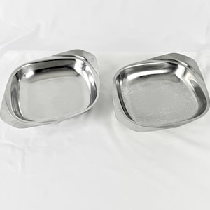 May include: Two stainless steel serving dishes. Each dish has a square shape with curved edges and a slightly raised rim. The dishes are made of shiny, reflective metal and are set against a white background.