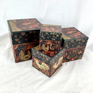 May include: Set of four square boxes with a green background and a colorful rooster design. The boxes are stacked on top of each other, with the largest box at the bottom and the smallest box at the top.