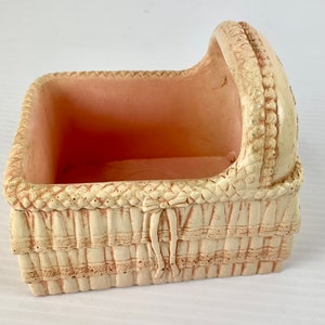 May include: A white ceramic baby crib with a pink interior. The crib has a woven design and a ruffled edge.