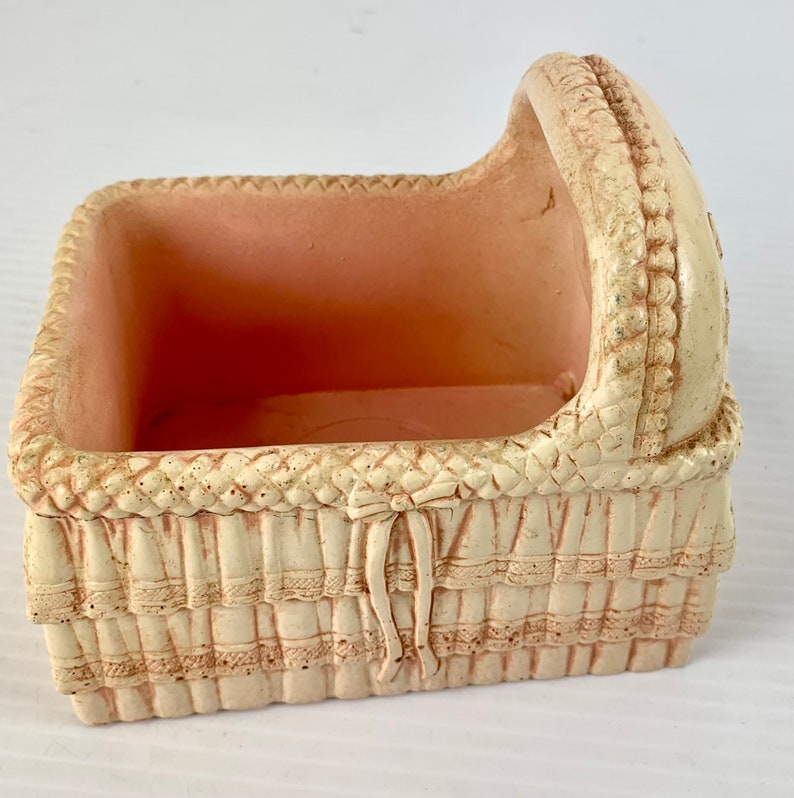 May include: A white ceramic baby crib with a pink interior. The crib has a woven design and a ruffled edge.