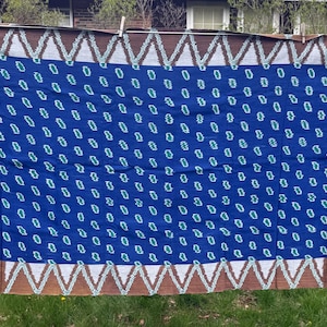 May include: A blue textile with a repeating white pattern and decorative borders. The borders feature red and white designs on the sides and a brown and white zigzag pattern along the top and bottom edges. The textile is hanging outside.