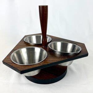 May include: A wooden condiment server with three stainless steel bowls. The server has a dark brown finish with a central wooden handle. The base is a dark brown circle with a black edge. The bowls are evenly spaced.