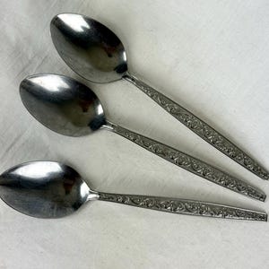 May include: Three stainless steel spoons with oval bowls and ornate, engraved handles. The spoons are arranged diagonally on a white surface, reflecting light. The handles feature a detailed floral pattern, adding a touch of elegance to the silverware.
