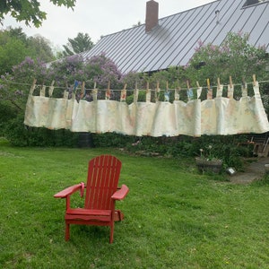 May include: A red Adirondack chair sits on a green lawn. A clothesline strung with floral-patterned fabric panels hangs in the background. The scene is set in a garden with a house and trees.