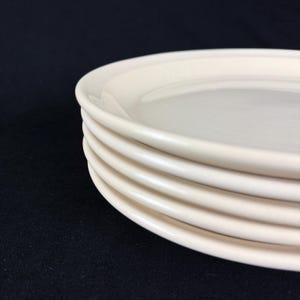 May include: A stack of off-white ceramic plates. The plates have a simple, classic design with a slightly raised rim. The plates are stacked neatly, with the edges of each plate visible. The background is a solid black.