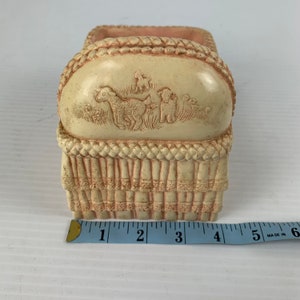 May include: A small, cream-colored ceramic crib with a pink woven design and a raised image of three lambs. The crib is approximately 3 inches wide.