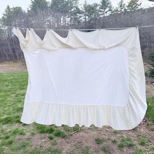 May include: A rectangular off-white bed sheet featuring a smooth central panel bordered by a textured, ruffled edge. The sheet is draped over a thin horizontal line, showcasing the gathered fabric along the top and the wide, pleated ruffle trim.