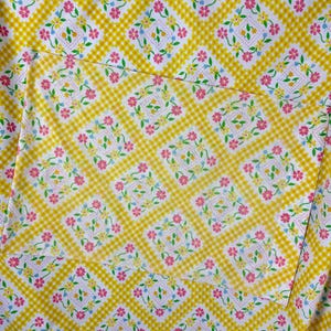 May include: A close-up of a fabric with a yellow gingham pattern overlaid with a floral design. The floral design features pink, yellow, and blue flowers arranged in a square pattern on a white background. The fabric has a pocket.