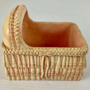 May include: A small, cream-colored ceramic planter shaped like a baby crib. The planter has a woven design and a raised bow on the front.
