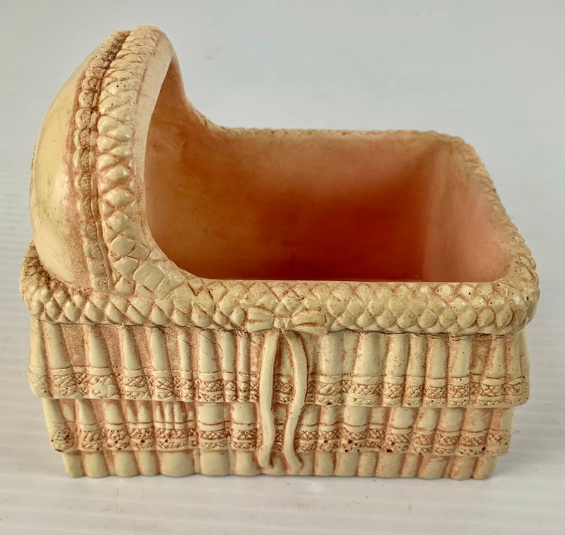 May include: A small, cream-colored ceramic planter shaped like a baby crib. The planter has a woven design and a raised bow on the front.