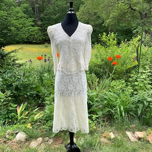May include: Ivory lace dress and jacket set displayed on a black mannequin. The dress has a midi-length skirt and the jacket has a V-neckline and button closure. The outfit is set against a backdrop of lush greenery and flowers.