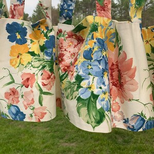 May include: Floral patterned curtains with a cream background. The curtains feature a variety of colorful flowers, including blue, yellow, pink, and red blooms, with green leaves. The curtains are hanging, with the top portion visible.