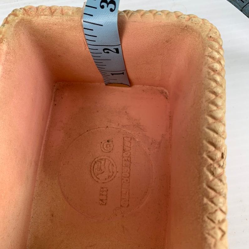 May include: A small, light pink, rectangular plastic container with a woven pattern around the edges. The container has a round, white label on the bottom with the text "Made in China" printed on it.