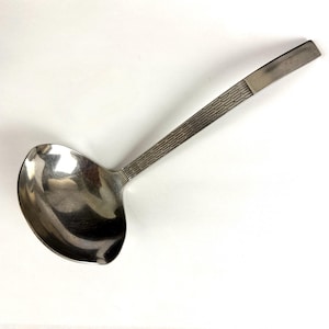 May include: A stainless steel soup ladle with a polished, rounded bowl and a long, slender handle. The handle features a repeating geometric pattern of horizontal lines and small vertical dashes, ending in a smooth, flat, rectangular tip.