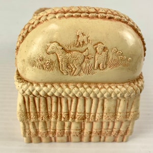 May include: A cream-colored ceramic baby crib with a rope-like trim and a raised design of three lambs on the top. The crib is a decorative piece.