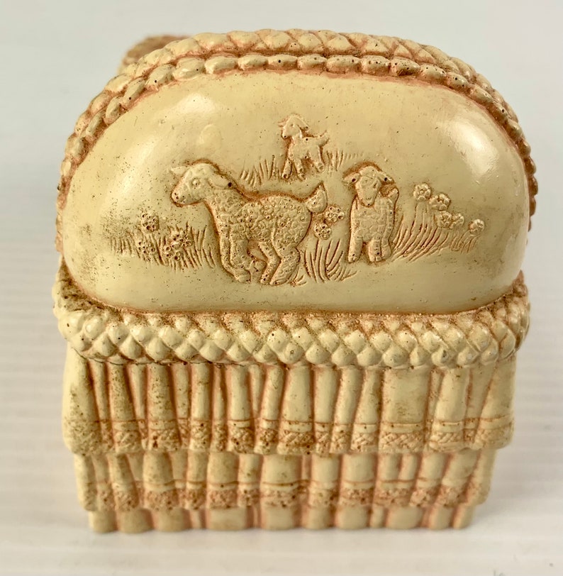 May include: A cream-colored ceramic baby crib with a rope-like trim and a raised design of three lambs on the top. The crib is a decorative piece.