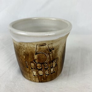 May include: A small, handmade ceramic cup with a brown, textured exterior and a white interior and rim. The words "5K Frosty Rock" are embossed on the side of the cup.