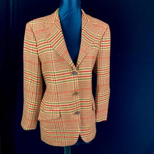 May include: A tailored blazer in a plaid pattern with red, green, and tan hues. The jacket features a notched lapel, three-button closure, and two front pockets. The blazer is displayed on a mannequin against a dark blue background.