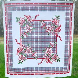 May include: A white tablecloth with a red and gray plaid pattern and a red border. The tablecloth features four floral arrangements with red flowers, green leaves, and red ribbons. The tablecloth is hanging outside on a clothesline.