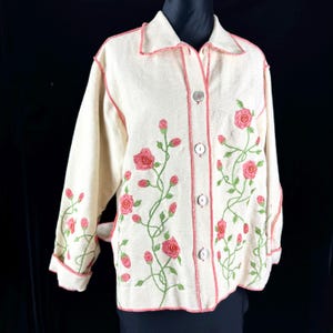 May include: Cream-colored button-up jacket with embroidered pink roses and green vines. The jacket has a collar, long sleeves, and pink stitching along the edges. The floral design is a prominent feature.