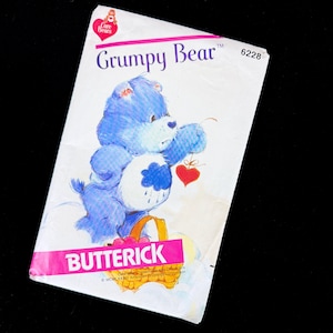 May include: A vintage Butterick pattern featuring the Care Bear character, Grumpy Bear. The pattern cover has a white background with a blue bear illustration holding a heart. The word "BUTTERICK" is printed in pink.