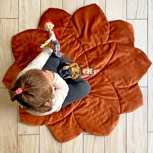 Könnte beinhalten: Ein Kind sitzt auf einer großen, braunen, blumenförmigen Spielmatte mit zwei Plüschtieren. Die Matte besteht aus weichem, samtigem Stoff und hat ein gestepptes Design.