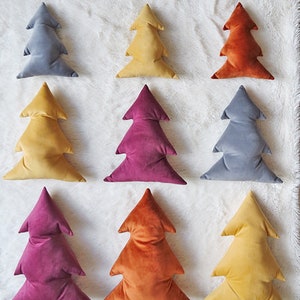 May include: Nine velvet Christmas tree pillows in various colors, including gray, yellow, orange, and burgundy. The pillows are arranged in three rows on a white fluffy surface. The text "OhOh deco" is visible in the bottom right corner of the image.
