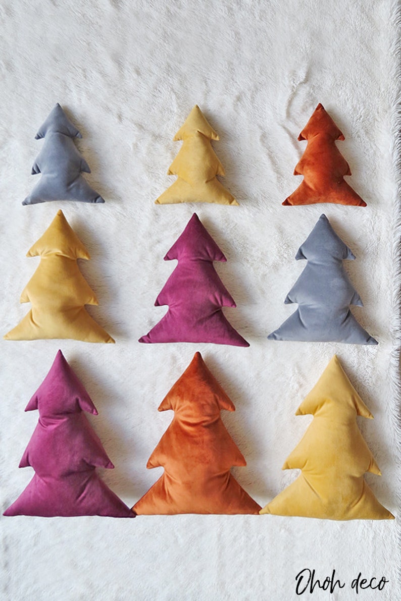 May include: Nine velvet Christmas tree pillows in various colors, including gray, yellow, orange, and burgundy. The pillows are arranged in three rows on a white fluffy surface. The text "OhOh deco" is visible in the bottom right corner of the image.