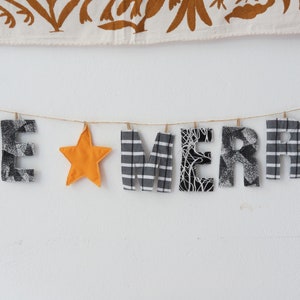 Peut inclure: Une bannière en tissu avec les mots "BE MERRY" en lettres de tissu noir et blanc. Une étoile jaune se trouve entre les mots.