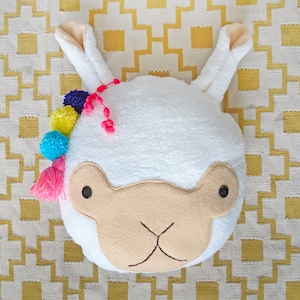 Puede incluir: Un cojín blanco con forma de llama con una superficie suave y texturizada. Tiene una cara beige, ojos negros de botón y una boca cosida. Pompones coloridos y una borla rosa adornan la parte superior. El fondo es un patrón geométrico amarillo y blanco.