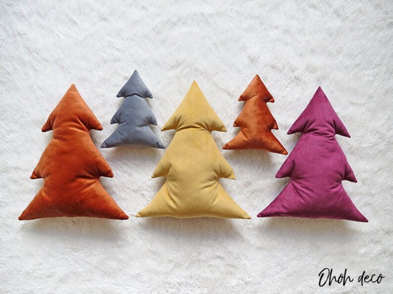 May include: Five velvet Christmas tree pillows in different colors: orange, gray, yellow, orange, and purple. The pillows are arranged in a row on a white fluffy surface. The text "OhOh deco" is visible in the bottom right corner of the image.