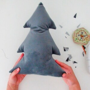 May include: A gray fabric Christmas tree pillow being held by two hands. The pillow is made of a soft, plush fabric and has a simple, classic design.