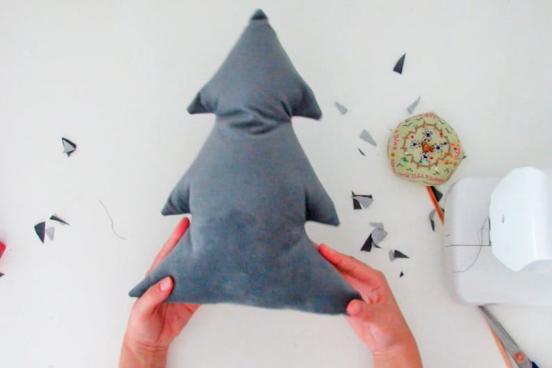 May include: A gray fabric Christmas tree pillow being held by two hands. The pillow is made of a soft, plush fabric and has a simple, classic design.