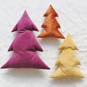 May include: Three velvet Christmas tree pillows in different sizes. The largest pillow is a deep purple, the medium pillow is a burnt orange, and the smallest pillow is a pale yellow.