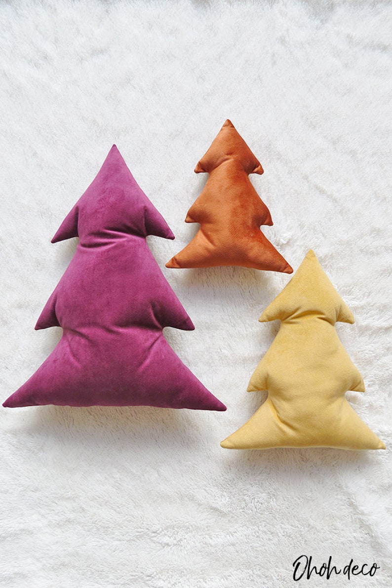 May include: Three velvet Christmas tree pillows in different sizes. The largest pillow is a deep purple, the medium pillow is a burnt orange, and the smallest pillow is a pale yellow.