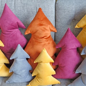 May include: A set of six plush velvet Christmas tree pillows in various colors, including orange, yellow, blue, and pink. The pillows are arranged in a row on a gray couch.