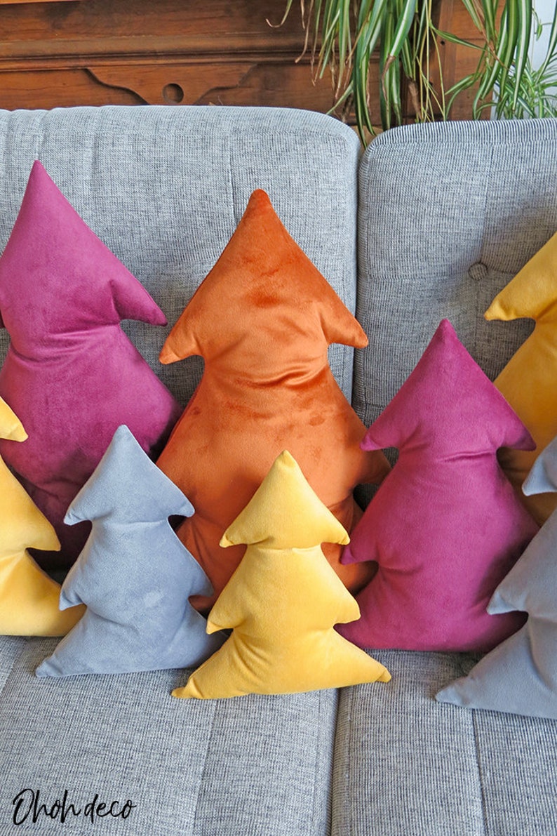 May include: A set of six plush velvet Christmas tree pillows in various colors, including orange, yellow, blue, and pink. The pillows are arranged in a row on a gray couch.