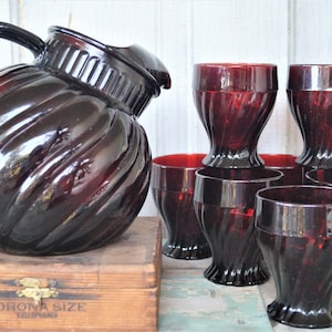 May include: A set of six ruby red glass tumblers and a matching pitcher with a swirl design. The pitcher has a handle and a wide mouth.