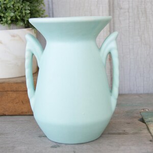 May include: A light blue ceramic vase with two handles. The vase has a wide mouth and a rounded body. The vase is sitting on a wooden surface. A small plant in a white pot is in the background.