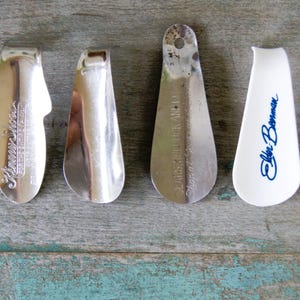 May include: A collection of six shoehorns in various colors and designs. The shoehorns are made of metal and plastic. Some have text, including "Florsheim", "Penny Moore", and "Elbe Bowman". The shoehorns are arranged on a weathered wooden surface.