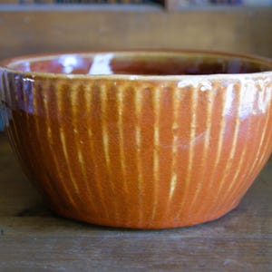 May include: A glazed, orange ceramic bowl with vertical ribbed detailing. The bowl has a rounded shape and a glossy finish, suitable for serving or display. The interior is a lighter shade of orange.