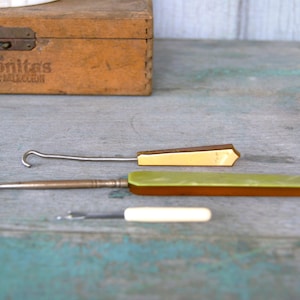 May include: A collection of vintage crochet hooks and tools, featuring metal hooks with wooden and plastic handles in shades of cream, green, and brown. A wooden box with the text "Coronitas Special Selection" is in the background.