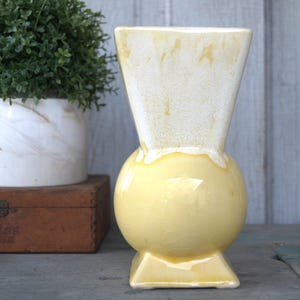 May include: A yellow and white ceramic vase with a rounded body and a square base. The vase has a unique, modern design.